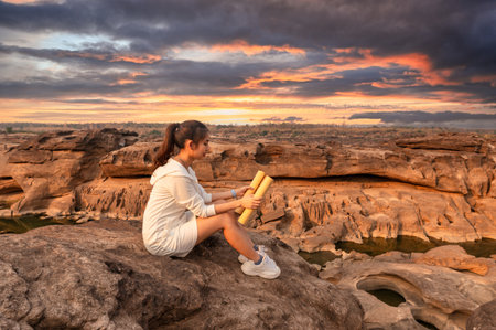 Beautiful young asian woman traveler sitting and looking at paper map on rock cliff in grand canyon of Thailand at sunsetの写真素材