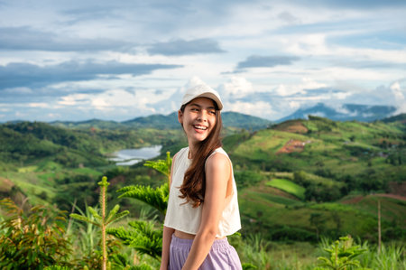 Happy asian woman wearing cap standing and smiling among the mountain with sunlight on vacationの写真素材