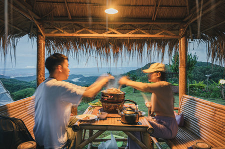 Young asian couple eating and grilling thai pork barbecue on pan in the hut along with a view of mist on hill in countrysideの写真素材