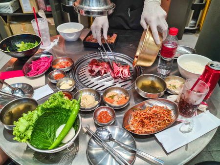 Staff is grilling sliced marinated pork on charcoal stove with korean side dishes and pickles in Korean restaurantの写真素材