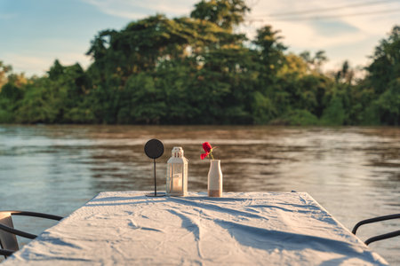 Flower vase, candle lantern, label on dining table with tablecloth at restaurant by riverfront in the sunsetの写真素材