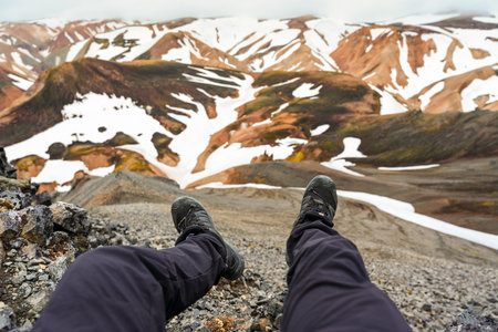 POV of a man with boots sitting and relaxing on the cliff among volcanic mountain on blahnjukur trail in summer at Landmannalaugar, Icelandの写真素材
