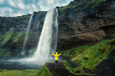 Cheerful asian woman in yellow jacket standing and looking at moody Seljalandsfoss waterfall flowing from cliff in summer at South of Icelandの写真素材
