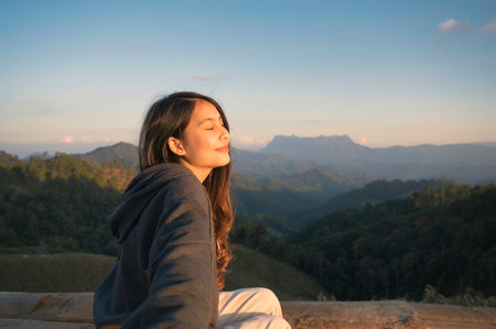 Beautiful young asian woman radiating happiness is sitting on top of mountain and warm sunshine in the evening at Doi Luang Chiang Dao national park, Thailandの写真素材