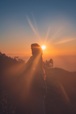 Young female wearing knit beanie  standing confrontation with sunrise on the mountain peak in tropical rainforest at national parkの写真素材