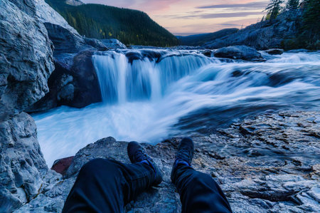POV legs of traveler sitting and relaxing beside rapids waterfall flowing in the forest during evening at Elbow falls, Kananaskis, Alberta, Canadaの写真素材