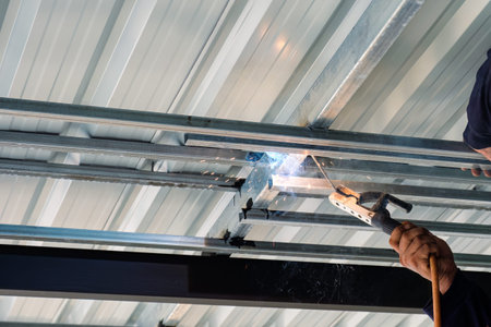 Construction worker installing and welding with flying sparks on steel frame of roof structure in...