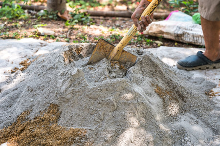 Construction worker manually mixes sand, cement, and water on the ground using a shovel and trowel,...