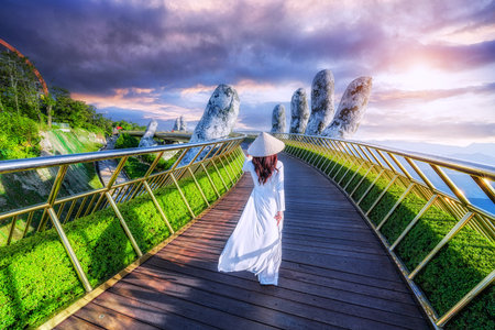 Vietnamese girl wearing white Vietnamese national costume walking on the golden bridge two giant hands on Ba Na Hill in Da Nang, Vietnam.の写真素材