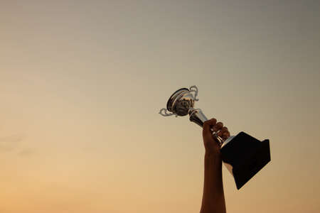 Victory concept with a hand holding a trophy on the sunset sky backgroundの写真素材
