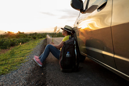 Woman sitting on the road at sunset With cars and backpacks Concept of vacation and travelの写真素材