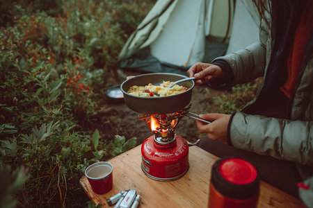 A close-up view of a person cooking a meal in a pan on a red portable gas stove. The stove is placed on a wooden surface, surrounded by lush green vegetation. Steam emanates from the pan, and a small flame is visible under it. A red cup and a thermos are also on the table, indicating a camping or outdoor cooking scenario.の写真素材