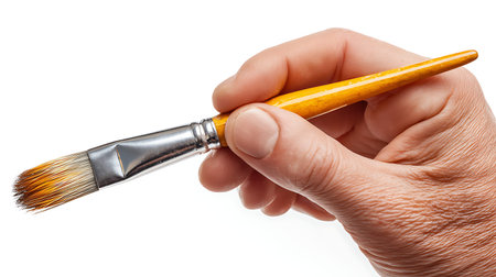 A detailed view of a human hand holding a paintbrush. The paintbrush has a smooth, yellow wooden...