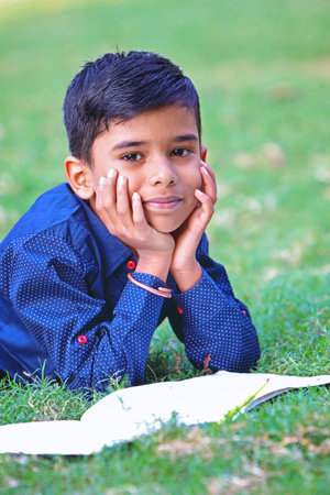Portrait of Indian little boy posing to camera, lying down on green grass with his text book