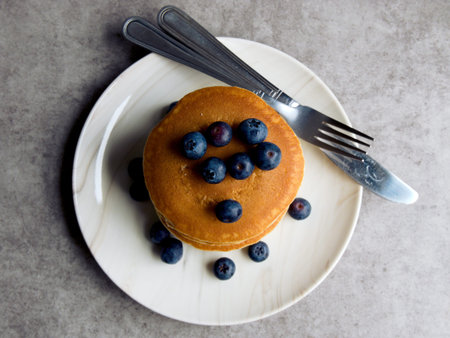 Pancakes with blueberries, stacked on a white plate with a knife and fork. Placed on a grey...