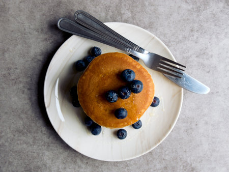 Pancakes with blueberries, stacked on a white plate with a knife and fork. Placed on a grey...
