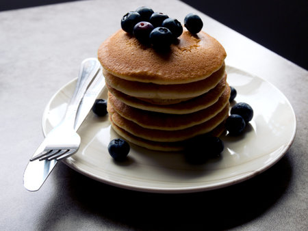 Pancakes with blueberries, stacked on a white plate with a knife and fork. Placed on a grey...