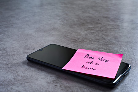 One step at a time sticky note left on smartphone. The bright note stands out against the keys,...