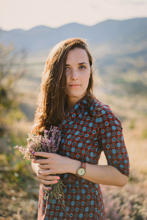 Young woman in patterned shirt standing and holding bouquet of wildflowers. Looking at camera with calm face. Sunny summer day.の写真素材