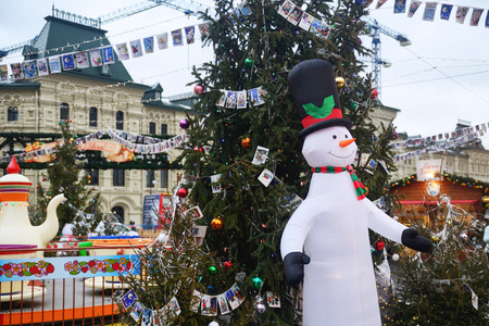 Funny snowman in a woolen scarf and hat on background of elegant Christmas fir in an amusement parkのeditorial素材