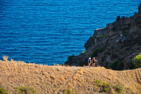 Couple of tourists are standing on a mountain top on a trail in the background of the blue seaの写真素材