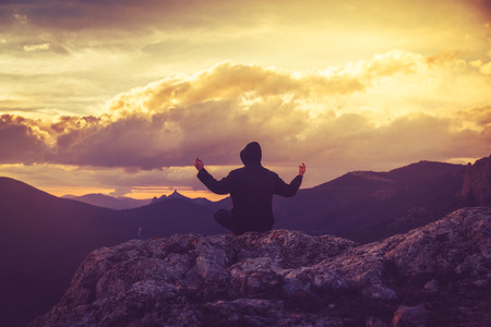 A young man is meditating in the lotus position on top of a mountain against the backdrop of a sunset, view from the backの写真素材