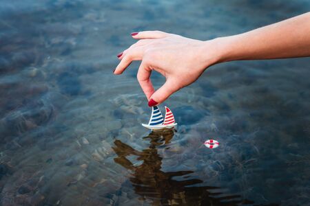 Small toy boat in a female hand above the water on the seashore, concept of vacation, vacation, tripの写真素材