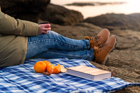 Girl on the beach on vacation on a plaid drinks tea, a book and tangerines in the front pallet,...