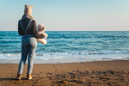 Girl with a teddy bear in hands on the seashore on a clear day, view of the backの写真素材
