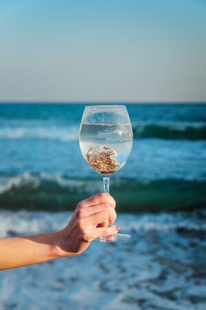 Seashells in a glass in a female hand on a background of blue seaの写真素材