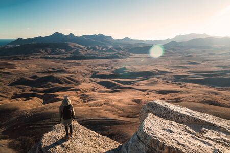 Girl with a backpack stands on the edge of a cliff in the mountains with a view of the valley at sunset, view from the back.の写真素材