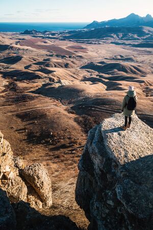 Girl with a backpack stands on the edge of a cliff in the mountains with a view of the valley at sunset, view from the back.の写真素材