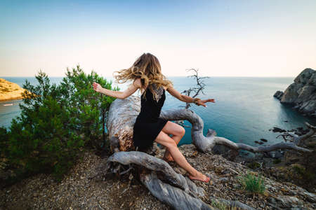 A girl in a black dress on the edge of a cliff against the background of the sea, long hair covers her face, quickly grows her head, fooling around. Beautiful summer landscapeの写真素材