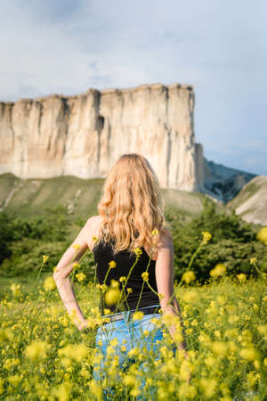 Blonde girl in a black bodysuit with a white hat walks in a rapeseed field, having fun, view from the backの写真素材