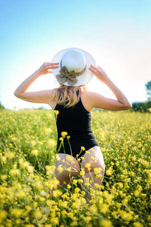 Blonde girl in a black bodysuit with a white hat walks in a rapeseed field, having fun, view from the backの写真素材