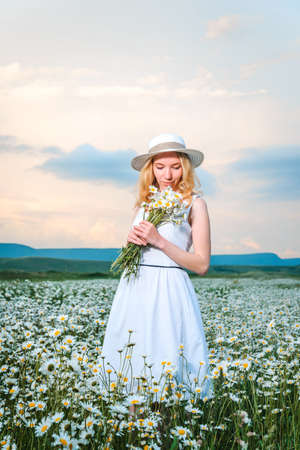 Girl in a hat and white dress with a bouquet of white wild daisies in a chamomile fieldの写真素材