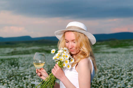 Girl in a hat and white dress with a bouquet of white wild daisies in a chamomile fieldの写真素材