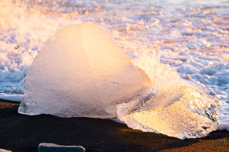 Pure glacier ice shining at sunset. Black volcanic sand at ocean coast. Pure ocean waves crash golden icebergs in Iceland. Diamond beach - famous tourist location in north Europe country.の写真素材