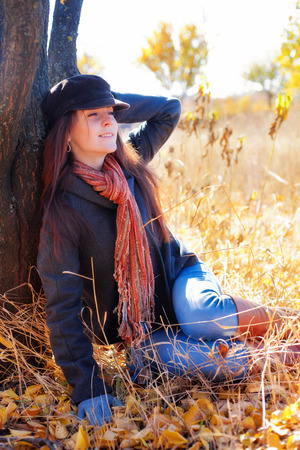Girl with smile is sitting near a tree on a sunny day in autumnの写真素材