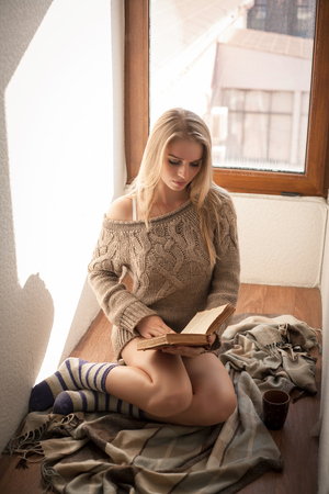 Young beautiful woman in a sweater reading a book near the window.の写真素材