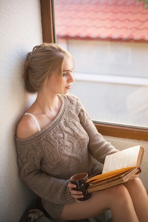 Young beautiful woman in a sweater reading a book near the window.の写真素材