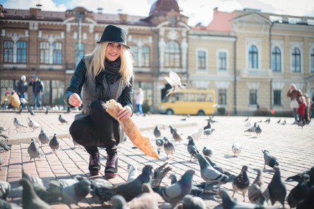 Young beautiful woman is feeding pigeons with bread crumbs in the town square on a sunny autumn dayの写真素材