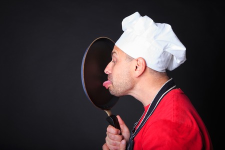 Studio shot of positive chef licks a frying pan, over dark background.の写真素材