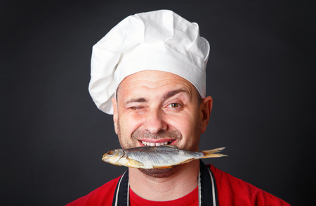 Funny cook in a hat with a dry fish in his teeth in the studio on a black background.の写真素材