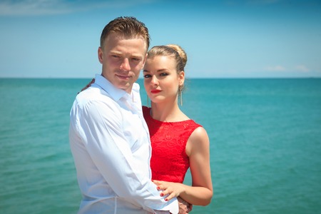Man is hugging a woman delicatly posing with her by the sea. Portrait of a beautiful couple on vacation.の写真素材