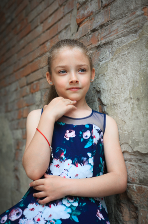 Portrait of a cute young school-age girl in a beautiful dress outdoors against a brick wall background.の写真素材