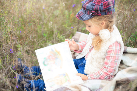 Little cute little girl is reading a book in nature against a background of autumn meadow.の写真素材