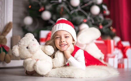 Lovely girl in santa hat on the floor near the Christmas tree with a bear, studio shot.の写真素材