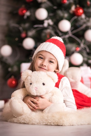 Lovely girl in santa hat sitting on the floor near the Christmas tree with a bear, studio shot, toning in vintage style.の写真素材