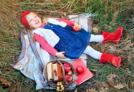 Little girl lying in grass on a plaid with a basket of fruit in the autumn day.の写真素材